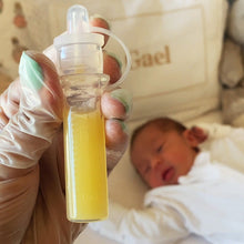 Load image into Gallery viewer, Haakaa colostrum collector filled with yellow liquid (colostrum breastmilk) held by a gloved hand, baby lying in crib in the background